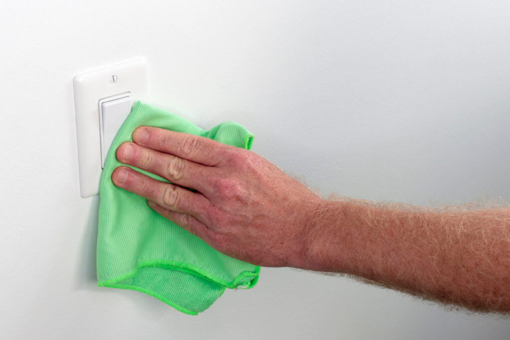 Persons hand wiping a wall white light control. Inside white wall light switch cleaned with a cloth. Indoor flat white light switch being cleaned by a hand with a rag as part of house cleaning chores.